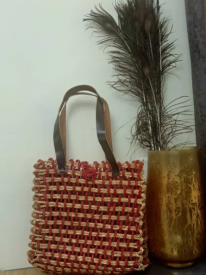 This tote bag features an open-weave design using a combination of natural and red-dyed fibers, creating a striking visual texture. Paired with dark leather-style straps, it’s a chic example of eco-friendly fashion.