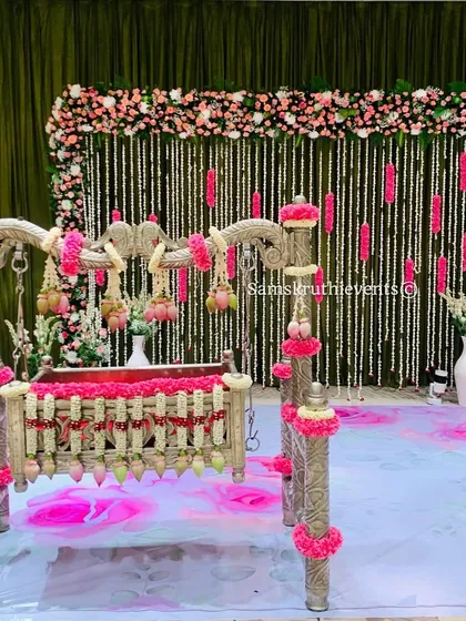 A modern naming ceremony setup with a silver cradle decorated with pink flowers. The backdrop features hanging jasmine strings against a green curtain, creating a fresh and elegant look.