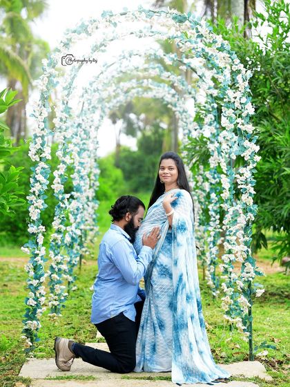 A romantic couple's portrait under a floral archway. The mother-to-be is wearing a tie-dye blue and white saree.