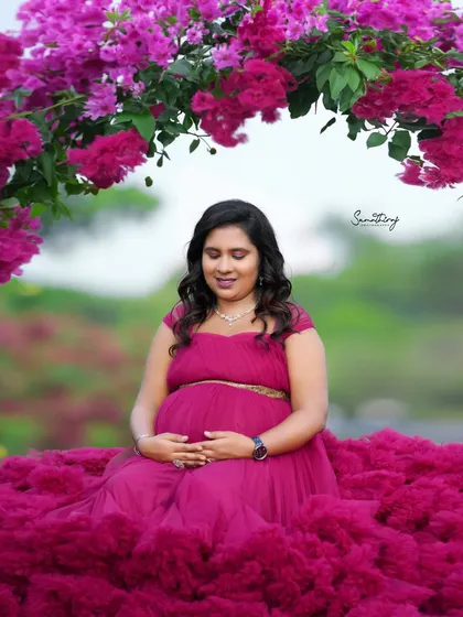 A serene outdoor portrait of a mother-to-be in a magenta gown, seated amidst a floral arch. Her peaceful expression and the beautiful surroundings create a magical moment.