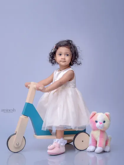 A little girl in a beautiful white dress poses with a modern wooden scooter and a colorful stuffed cat. The clean, light purple background creates a soft and dreamy feel.