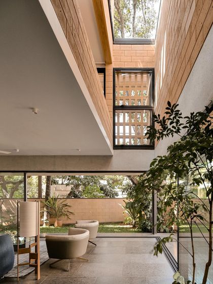 In the Debris Block House, a double-height space with a large skylight brings the outdoors in. The design uses jaali screens and large windows to connect the interior with the surrounding trees and garden.