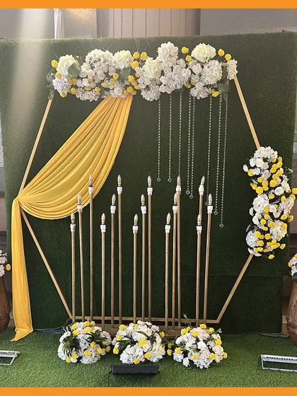 A modern and elegant photo corner for a Mehendi, with a hexagonal frame, yellow drapes, and tall candle-like fixtures.