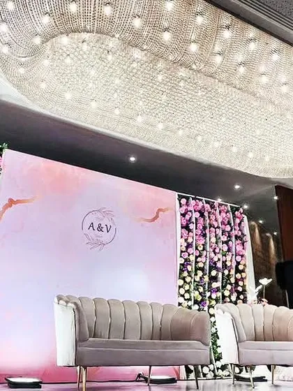 A sophisticated reception stage for 'A & V' under a stunning crystal chandelier. The design features a soft pastel backdrop flanked by walls of beautiful purple and pink flowers, creating a look of modern luxury.
