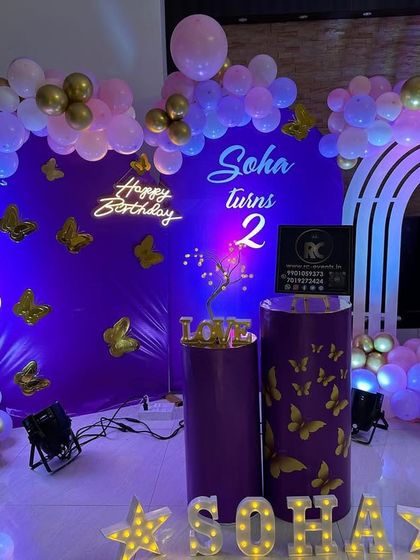 An elegant butterfly-themed setup in deep purple with gold accents. The decor features a neon sign, an arched backdrop, and marquee letters, perfect for a stylish second birthday.
