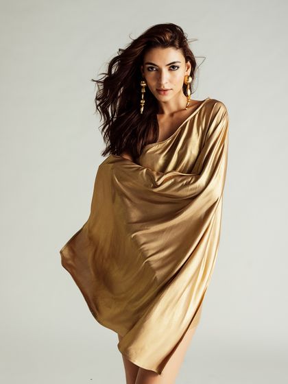 A studio shot featuring a flowing, one-shoulder gold dress. The styling is glamorous and goddess-like, with the wind machine used to create movement and drama.
