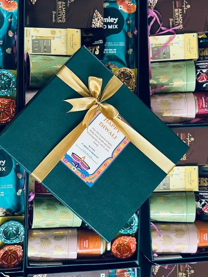 Celebrate in style with our elegant festive hampers. This custom Diwali gift for South Western Railways features a deep green box tied with a gold ribbon and a personalized "Happy Diwali" tag, surrounding a curated selection of treats.
