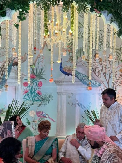 A candid moment from a wedding ceremony, with the couple seated against a stunning Pichwai art backdrop, surrounded by family.