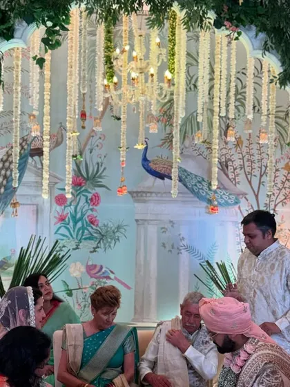 A candid moment from a wedding ceremony, with the couple seated against a stunning Pichwai art backdrop, surrounded by family.