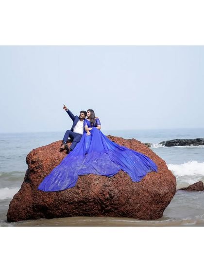 A wide shot of a couple on a large rock by the sea. The long, sparkling trail of the blue gown creates a magnificent, flowing look.