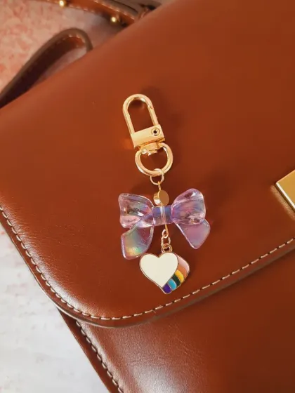 This bag charm combines an iridescent purple bow with a colorful rainbow heart charm, creating a magical and eye-catching accessory.