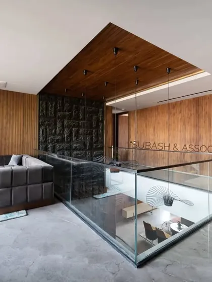This family lounge on the upper floor is surrounded by veneer-clad walls, concealing bedroom doors to create a clean, cohesive look. A glass railing maintains a view of the floor below.
