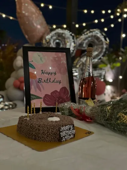 The essential birthday setup: a delicious cake, a bottle of bubbly, and a bouquet of flowers, all complemented by a personalized birthday card and festive balloons in the background.