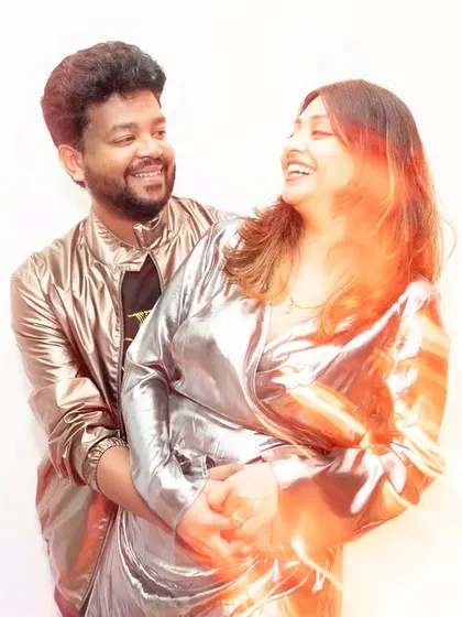 A joyful, candid moment captured with a warm light flare effect. The couple laughs together, their shiny outfits catching the light and adding to the photo's dynamic, celebratory energy.