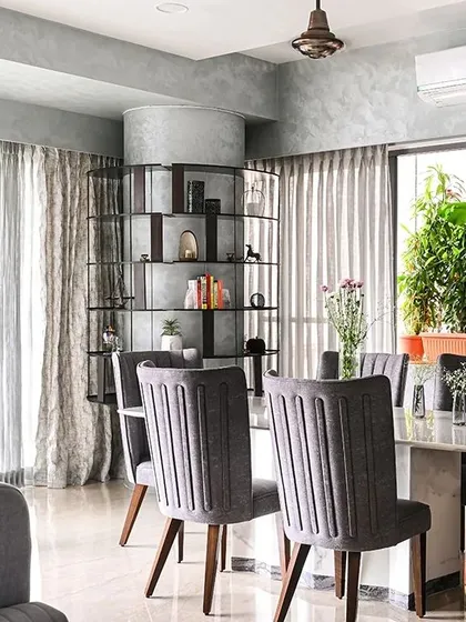 The dining area is defined by a marble-topped table and elegant grey upholstered chairs. In the background, the custom-built circular shelf around the column acts as a functional art piece, separating the dining space without closing it off.