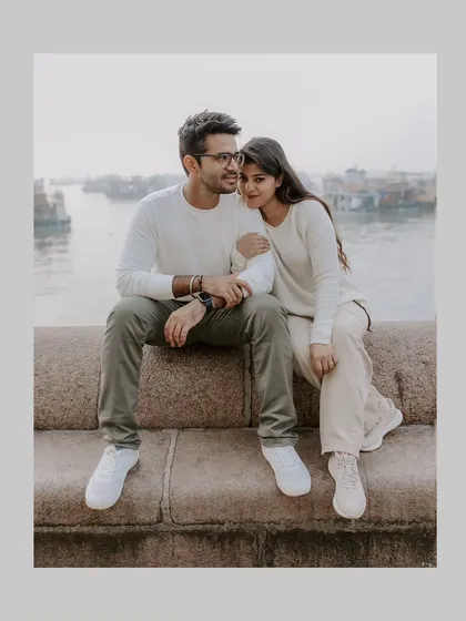 A quiet, intimate portrait of a couple sitting by the water near the Gateway of India, with the bustling Mumbai harbor in the background.