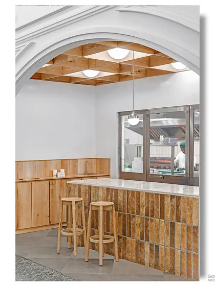 An interior shot of the cafe, focusing on the wooden bar and stools. The design is simple and modern, with a clear view into the kitchen, suggesting transparency and freshness.