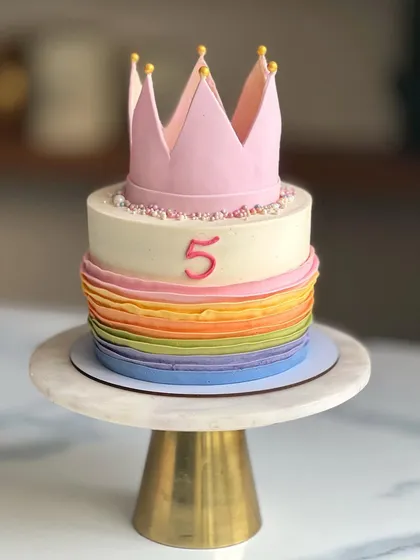 A princess cake with a modern twist. The bottom tier features colorful buttercream ruffles, while the top is kept simple and elegant, finished with a large pink fondant crown.