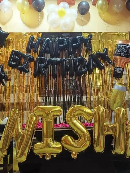 A fun and vibrant home birthday setup in black, gold, and yellow. The decor includes a foil curtain backdrop, name balloons, and fun foil balloons like a whiskey bottle, perfect for an adult's birthday party at home.