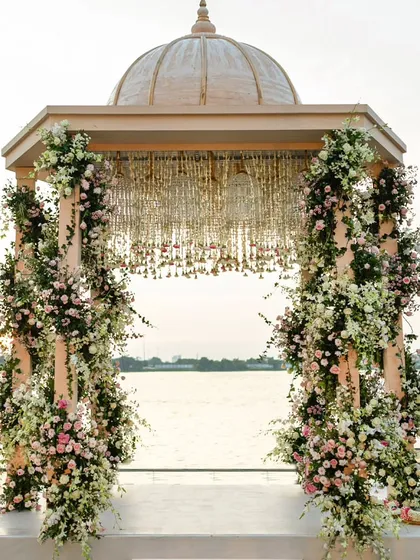 A picturesque Varmaala mandap set against the serene backwaters at sunset. The structure is adorned with spiraling garlands of pink and white roses, and the ceiling features a chandelier of cascading jasmine.