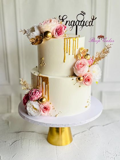 An elegant two-tier engagement cake designed with white frosting, a luxurious gold drip, and arrangements of pink and white roses. This cake is perfect for celebrating a promise of forever.