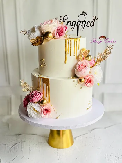 An elegant two-tier engagement cake designed with white frosting, a luxurious gold drip, and arrangements of pink and white roses. This cake is perfect for celebrating a promise of forever.