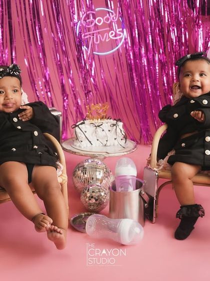 These twin divas are ready for their half-birthday party, sitting in tiny chairs with a cake and bottles ready for a celebration.