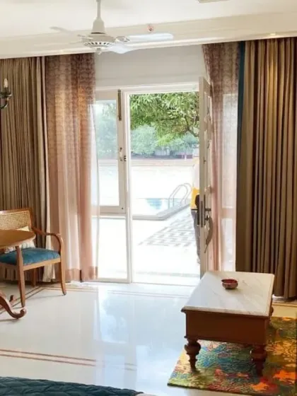 A living area in a suite at IHCL Pilibhit House, opening onto a poolside view. The design is clean and classic, with comfortable seating and rich brown curtains that frame the beautiful outdoor scenery.