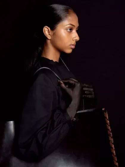 A moody, atmospheric shot from the Chamar Studio campaign at LFW. The makeup is minimal and understated, with a focus on sleek, wet-look hair to create a dramatic, high-fashion silhouette against the dark background.