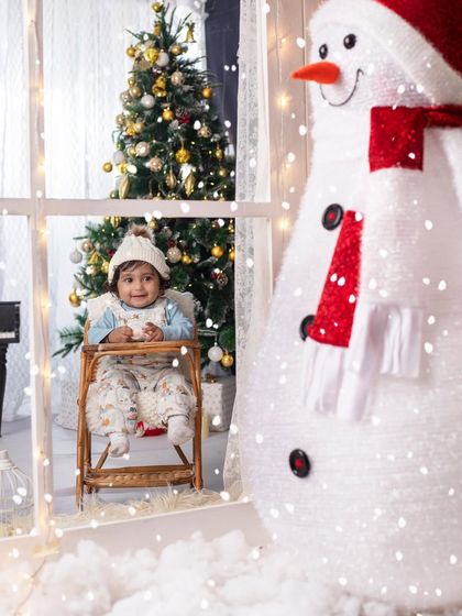 A little one sits by the window, watching the snow fall, with a happy snowman looking on. A perfect picture of winter magic.