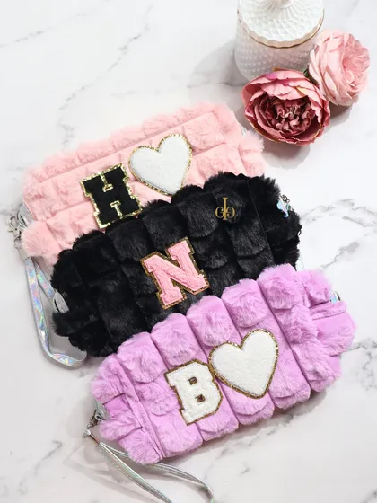 A collection of our fluffy fur pouches in pink, black, and lilac. They can be customized with multiple chenille patches, like initials and hearts, for a fun, personalized look.