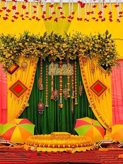 This is a full stage setup for a Haldi ceremony on a red carpet. The backdrop uses green and yellow drapes with intricate hangings, and the stage is set with a traditional 'urlī' and colorful umbrellas.
