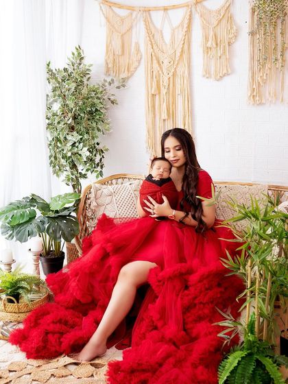 A mother in a vibrant red gown holds her baby, creating a striking and beautiful portrait in a bohemian-styled studio setting.