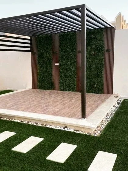 A completed pergola with a metal frame, a tiled floor, and a back wall featuring alternating panels of wood and artificial greenery. A stone paver path leads to the structure across an artificial lawn.