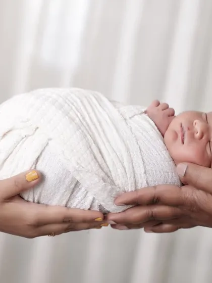 The delicate beginning of a new life. This iconic newborn photo shows a peacefully sleeping baby, swaddled in white, held securely in the loving hands of both parents. It's a simple, powerful shot that symbolizes family and protection.