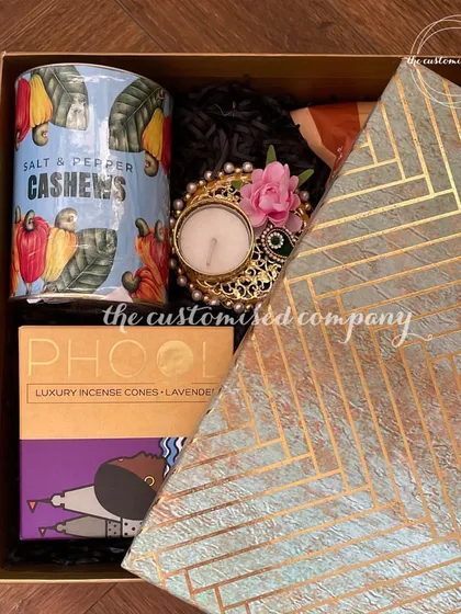 A peek inside a corporate gift box, featuring a mix of treats and tradition. It includes a can of flavored cashews, lavender incense cones from Phool, and a beautiful floral diya for the festive season.
