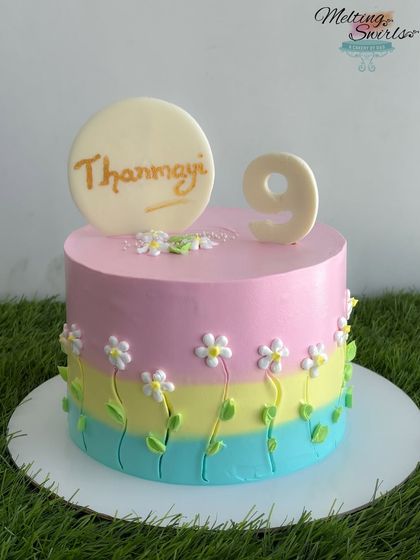 A pastel dream, this cake features pink, yellow, and blue stripes decorated with hand-piped daisies. The name and number are crafted from white chocolate.