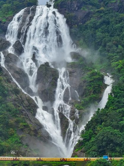A wide-angle view showing the entire expanse of Dudhsagar Falls with a train passing by, a truly bucket-list experience.