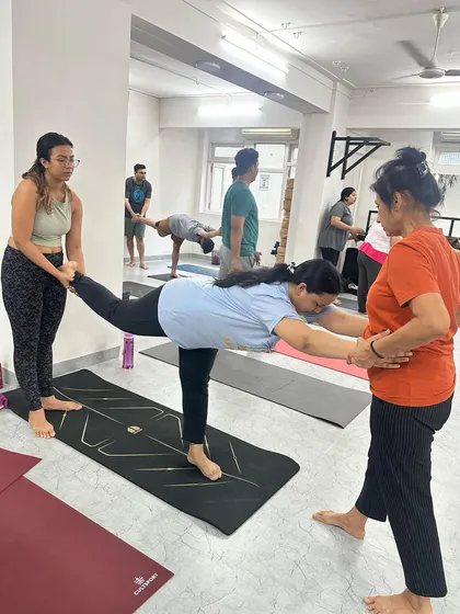 An instructor and a fellow student assist a practitioner in Virabhadrasana III (Warrior III), providing a stable frame for her to find her balance.