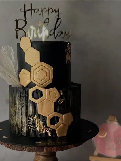For a bold and modern statement, this two-tier black fondant cake features a striking gold honeycomb design and a wood-grain texture on the bottom tier. A single white feather adds a touch of softness to this contemporary piece.