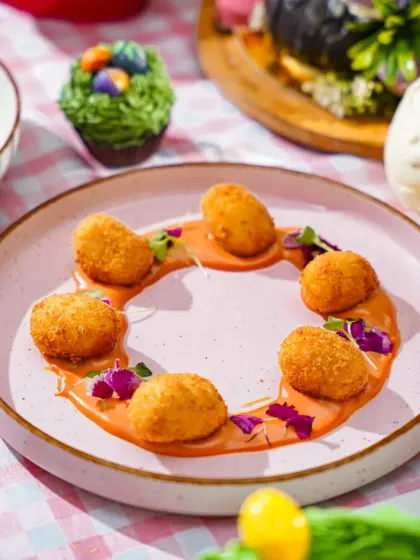 A plate of golden-brown croquettes arranged in a circle over a savory sauce, part of a creative Easter brunch. The bright, festive table setting adds to the celebratory mood.