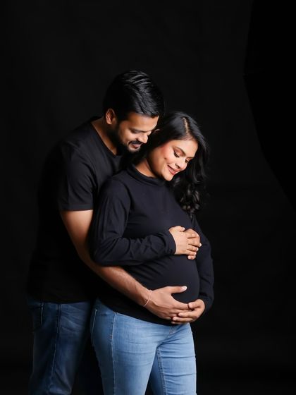 A casual and intimate maternity portrait in black and white. The couple is dressed in simple, dark clothes, putting all the focus on their connection and the baby bump.