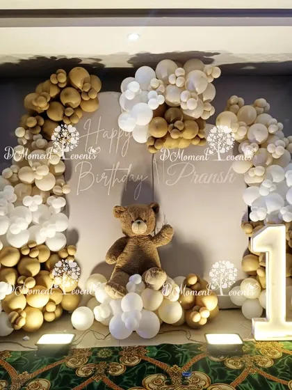 A cozy and cute teddy bear-themed first birthday backdrop. A large teddy bear sits among clouds of white and brown balloons, with a "Happy Birthday" sign and a "1" marquee light.