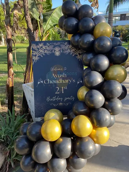 A personalized welcome sign for Ayush's 21st birthday party, framed by a black and gold balloon garland.