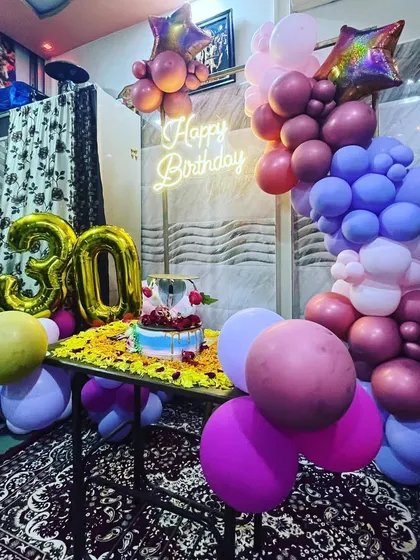 A cozy 30th birthday celebration at home. The decor includes a colorful balloon garland, a neon sign, and a large foil number '30' set up around the cake table.