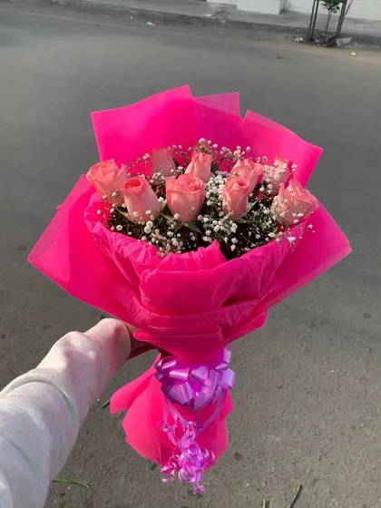 A vibrant bouquet of pink roses and baby's breath held outdoors. The bright pink wrapping paper makes this a cheerful and eye-catching gift.