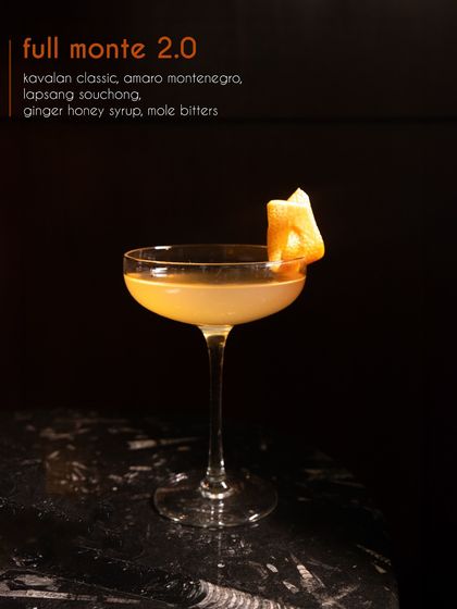 A different perspective on the Full Monte 2.0. This image highlights the cocktail's beautiful amber color and the elegant orange peel garnish, showcasing the balance of smoky lapsang souchong and ginger honey syrup.
