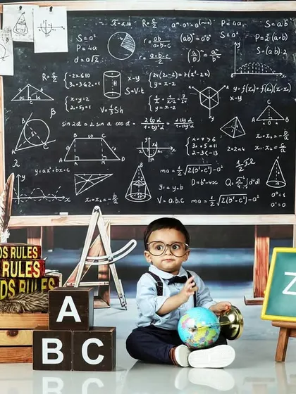 A "little genius" themed shoot, with a baby boy in glasses sitting in a classroom setup инфекции a chalkboard full of equations.