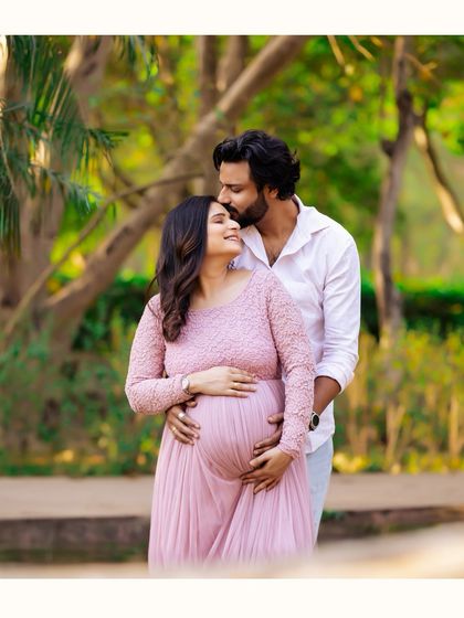 A tender embrace from behind, with the partner gently holding the baby bump. The soft light and natural park background make this a warm and intimate portrait.