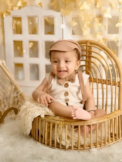 A smiling baby boy sits in a miniature wicker peacock chair, part of a warm and earthy boho-themed studio session.